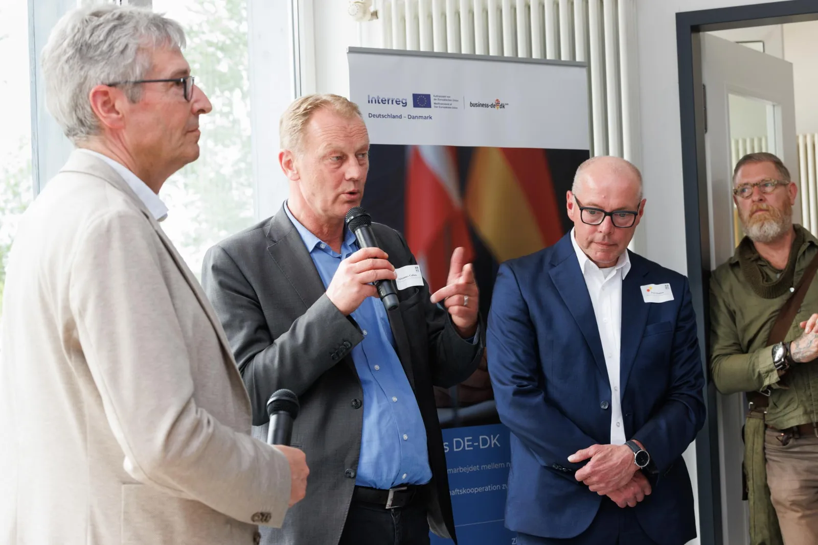 Tre mænd ved et Business DE-DK-community-møde, hvor den midterste taler i mikrofon foran et Interreg-banner
